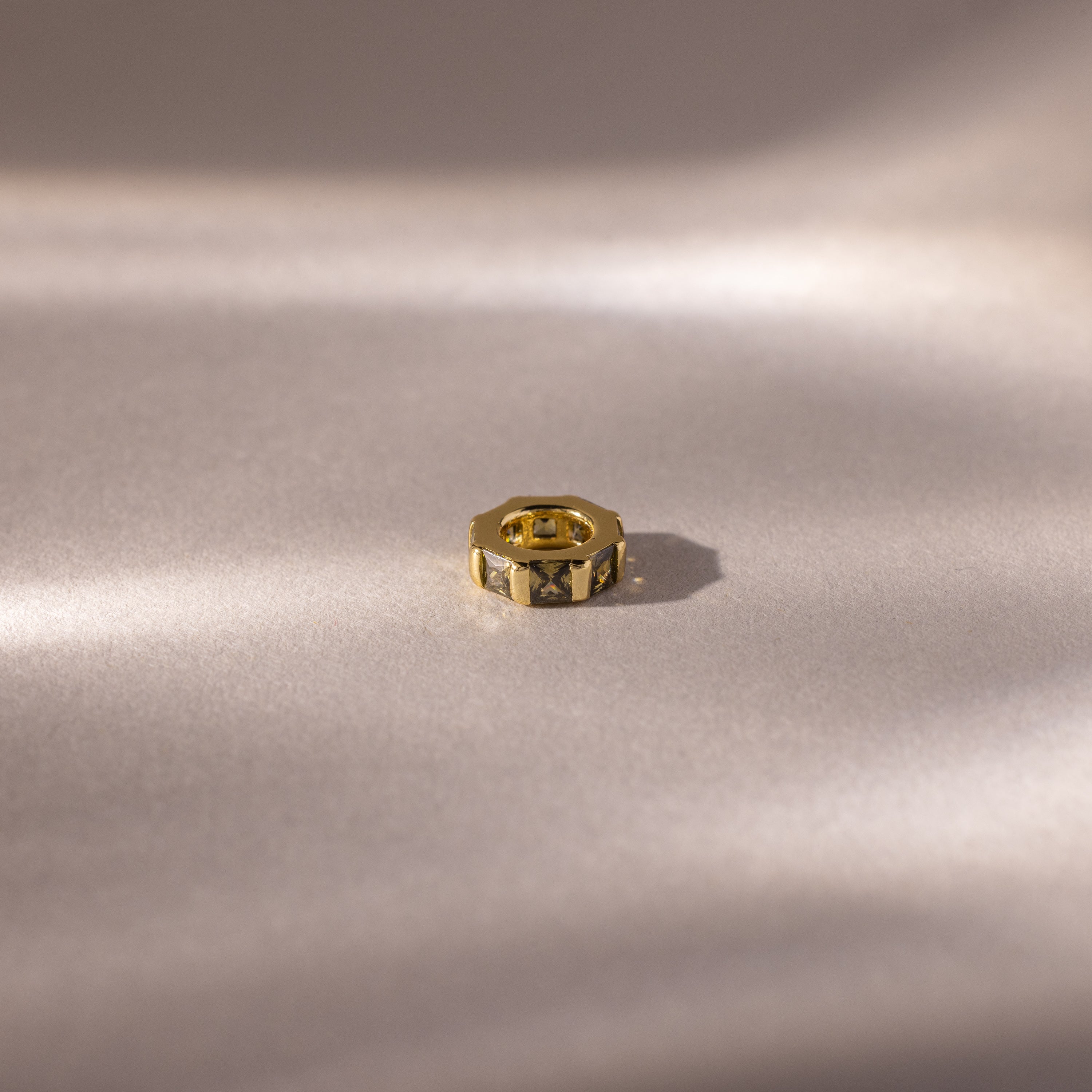 A small, gold hexagonal ring with an Infinity Princess Peridot Charm rests on a smooth, softly lit beige surface.