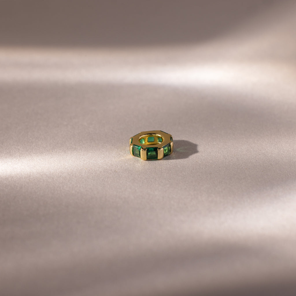 The Infinity Princess Emerald Charm, a gold ring with green princess-cut stones, rests on a soft, light beige surface with gentle shadows.