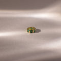 The Infinity Princess Emerald Charm, a gold ring with green princess-cut stones, rests on a soft, light beige surface with gentle shadows.