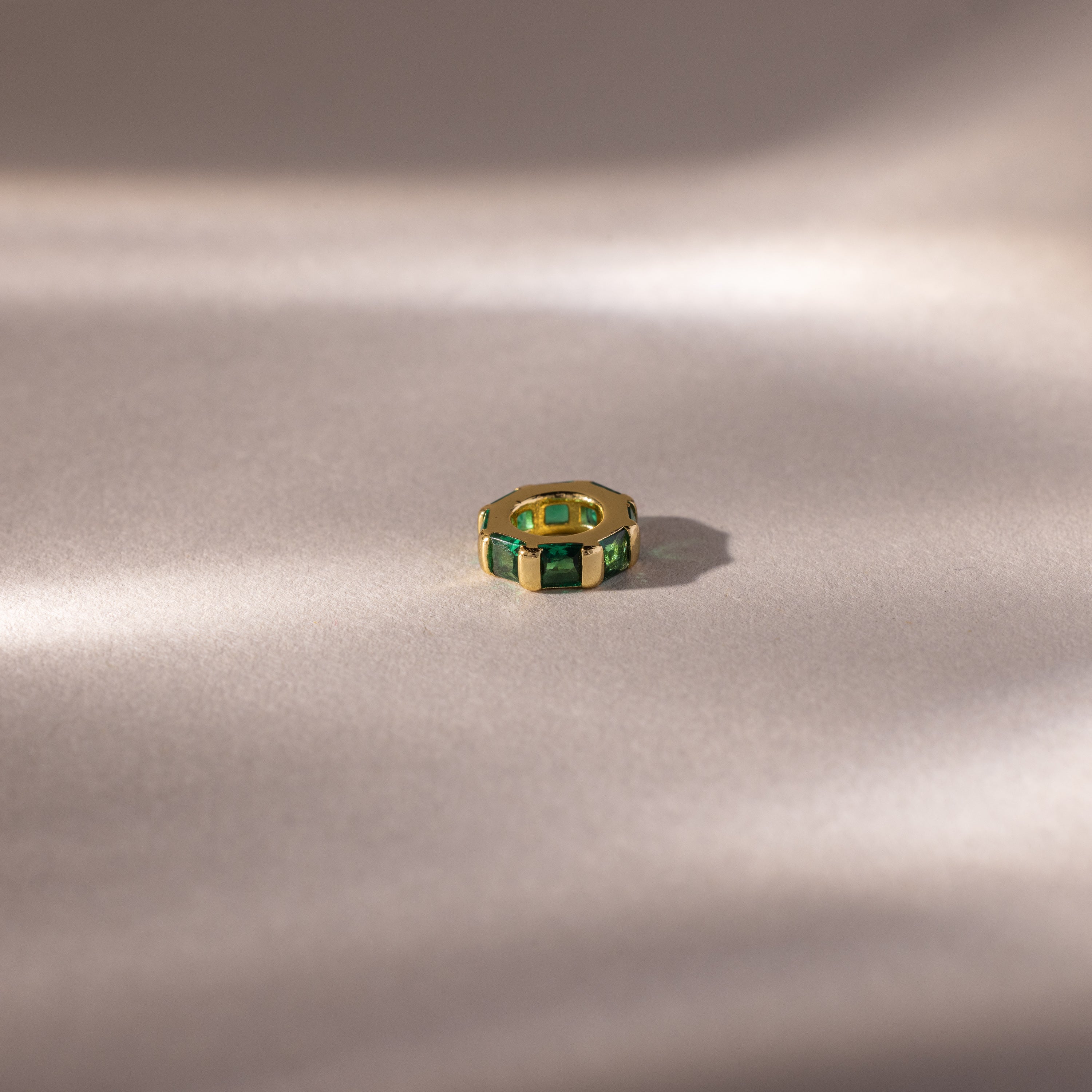 The Infinity Princess Emerald Charm, a gold ring with green princess-cut stones, rests on a soft, light beige surface with gentle shadows.