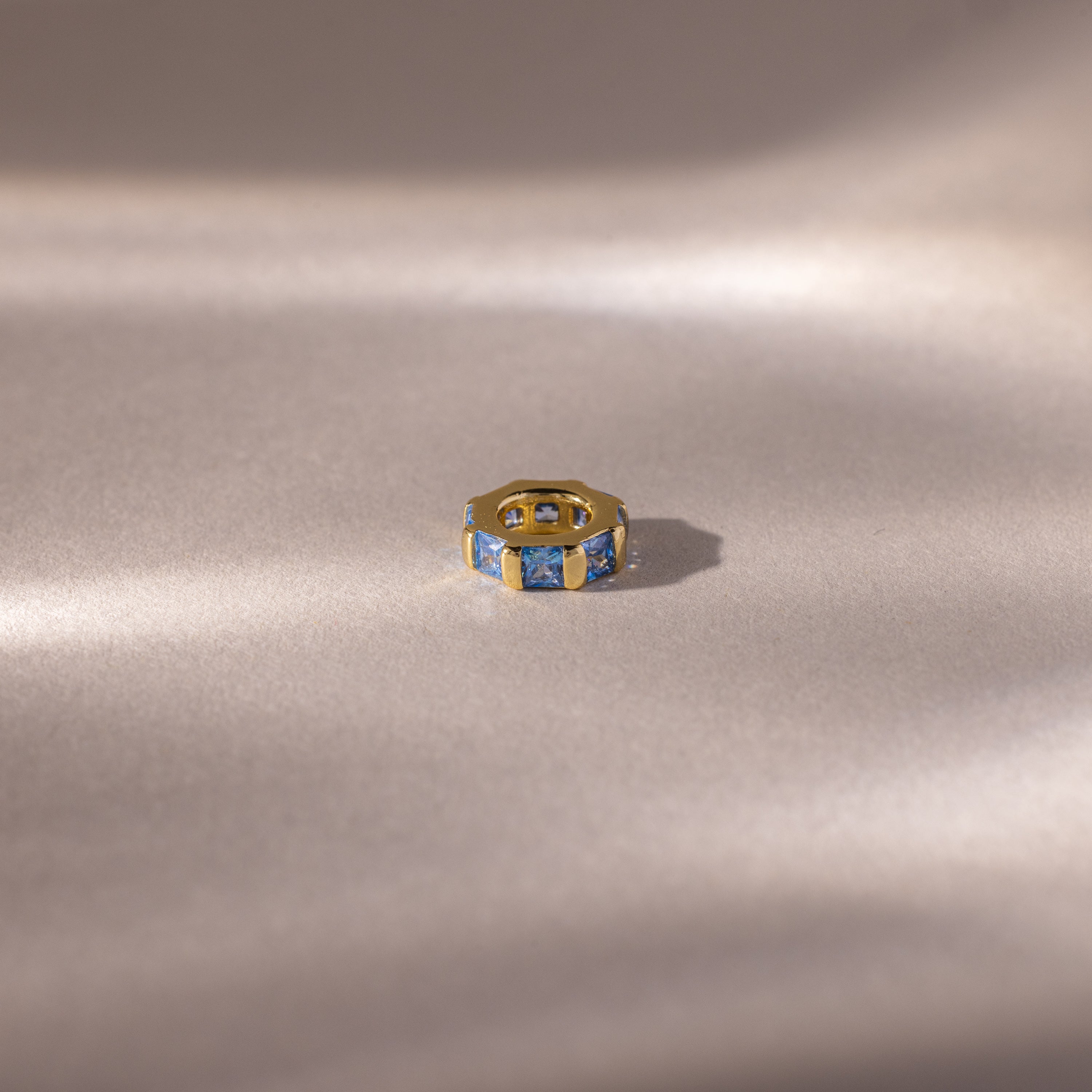 The Infinity Princess Blue Zircon Charm, featuring princess-cut blue stones, is elegantly displayed on a soft beige surface with gentle lighting—a stylish tribute to timeless design.