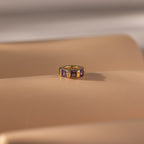The Infinity Princess Birthstone Charm features princess-cut purple stones, showcased on a smooth beige surface.