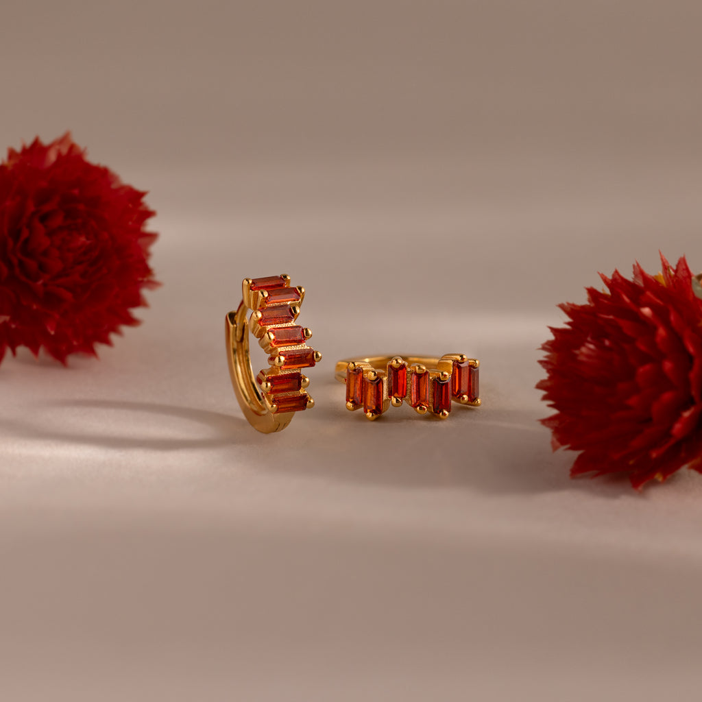 Staggered Baguette Birthstone Huggies with red baguette-cut stones are displayed between two red flowers on a neutral background, highlighting their modern elegance.