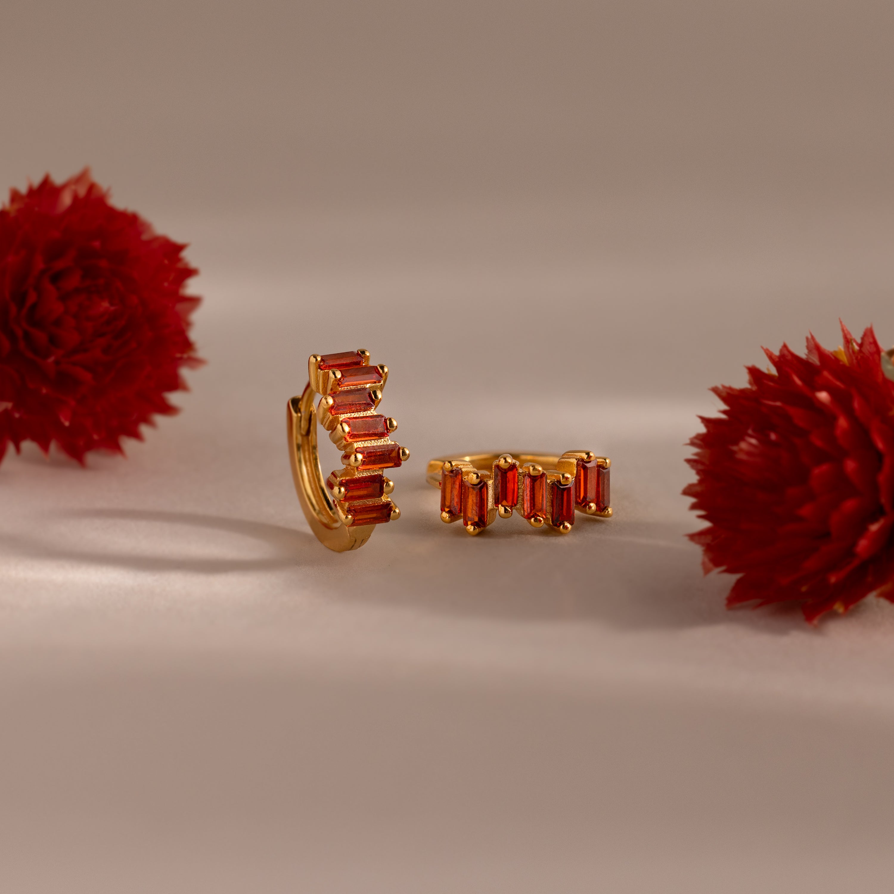Staggered Baguette Birthstone Huggies with red baguette-cut stones are displayed between two red flowers on a neutral background, highlighting their modern elegance.