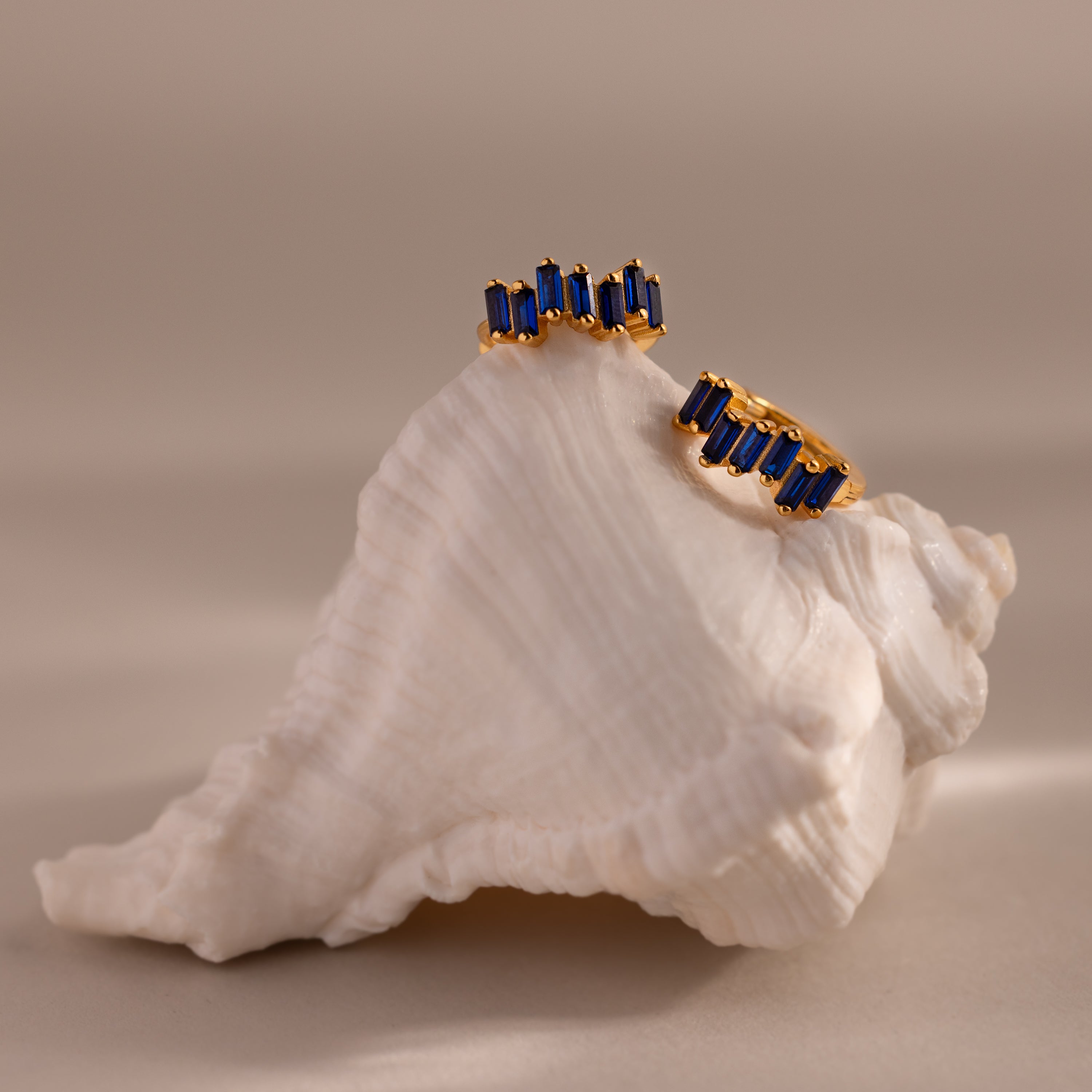The Staggered Baguette Birthstone Huggies, featuring blue baguette-cut stones, are elegantly displayed on a white seashell against a soft, neutral background.
