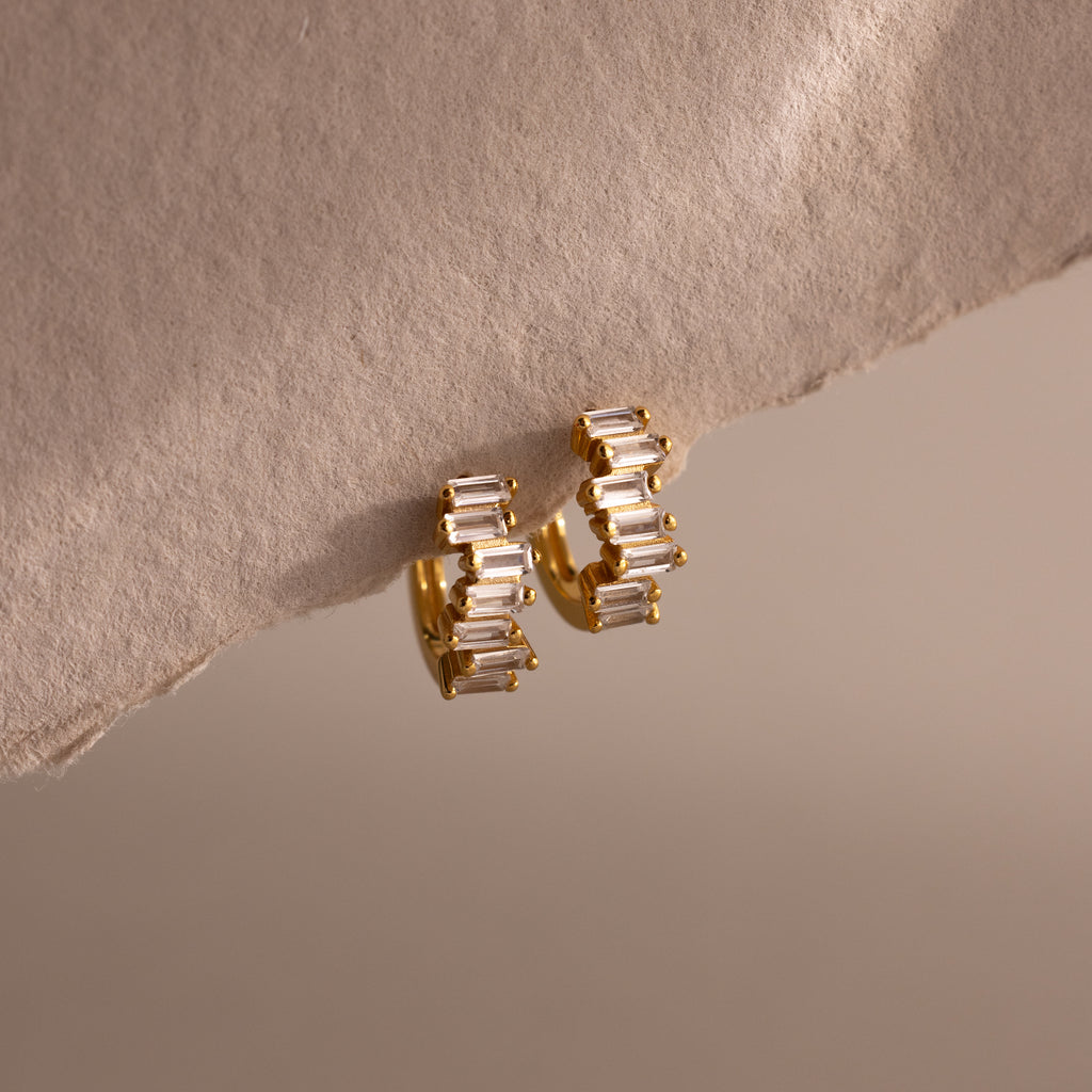 Staggered Baguette Diamond Huggies, featuring gold modern earrings with baguette-cut stones, shown on a textured beige surface with soft lighting.