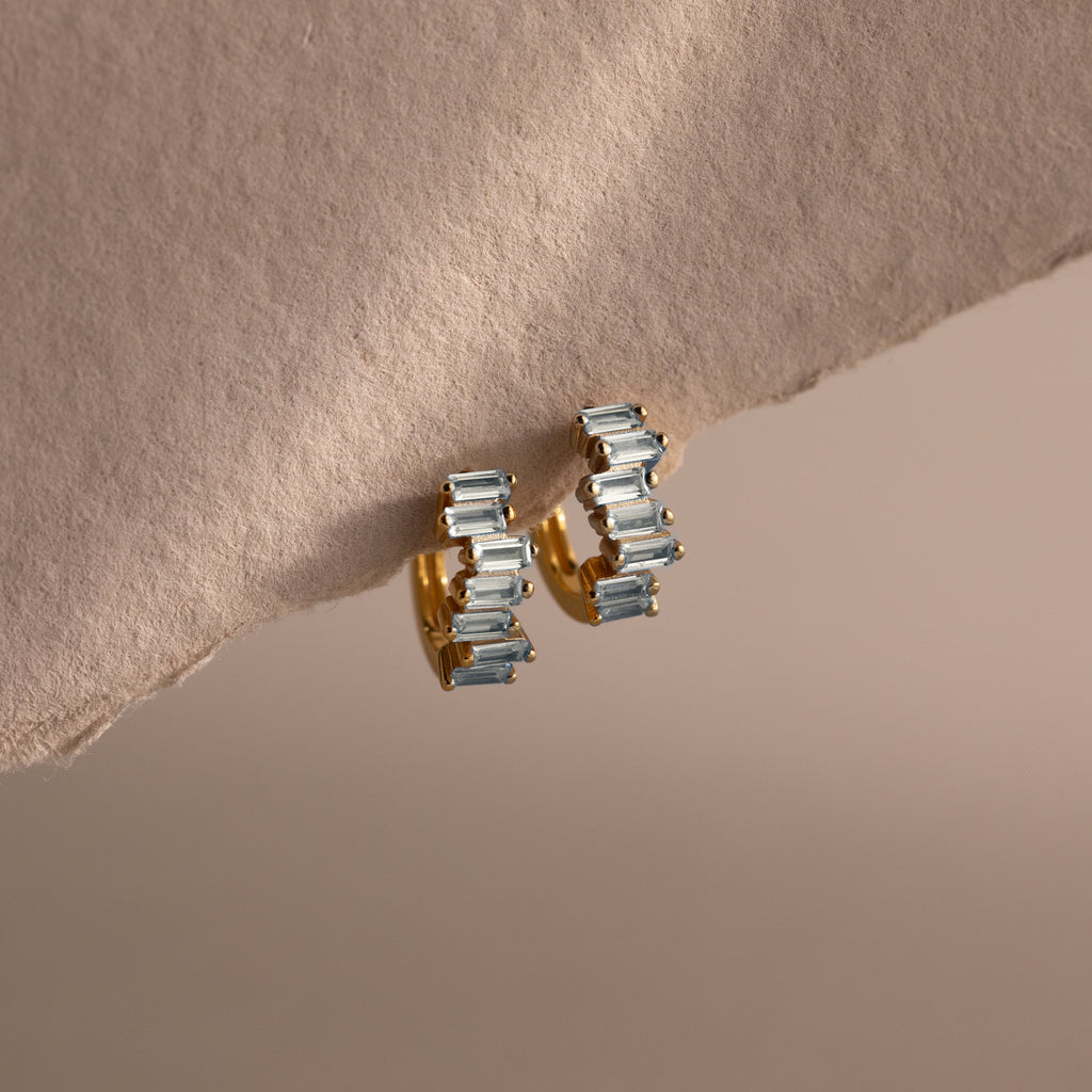 Staggered Baguette Aquamarine Huggies featuring rectangular aquamarine gemstones are shown on a soft, beige textured surface.