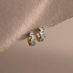 Staggered Baguette Aquamarine Huggies featuring rectangular aquamarine gemstones are shown on a soft, beige textured surface.