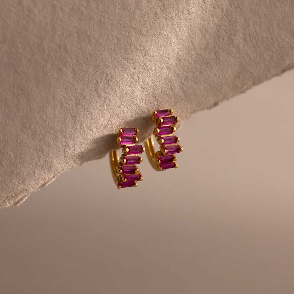 Staggered Baguette Ruby Huggies, featuring gold hoops with rectangular pink baguette-cut stones, are displayed on a textured neutral surface under soft lighting for a modern, elegant touch.