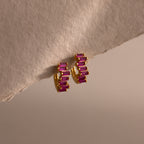 Staggered Baguette Ruby Huggies, featuring gold hoops with rectangular pink baguette-cut stones, are displayed on a textured neutral surface under soft lighting for a modern, elegant touch.