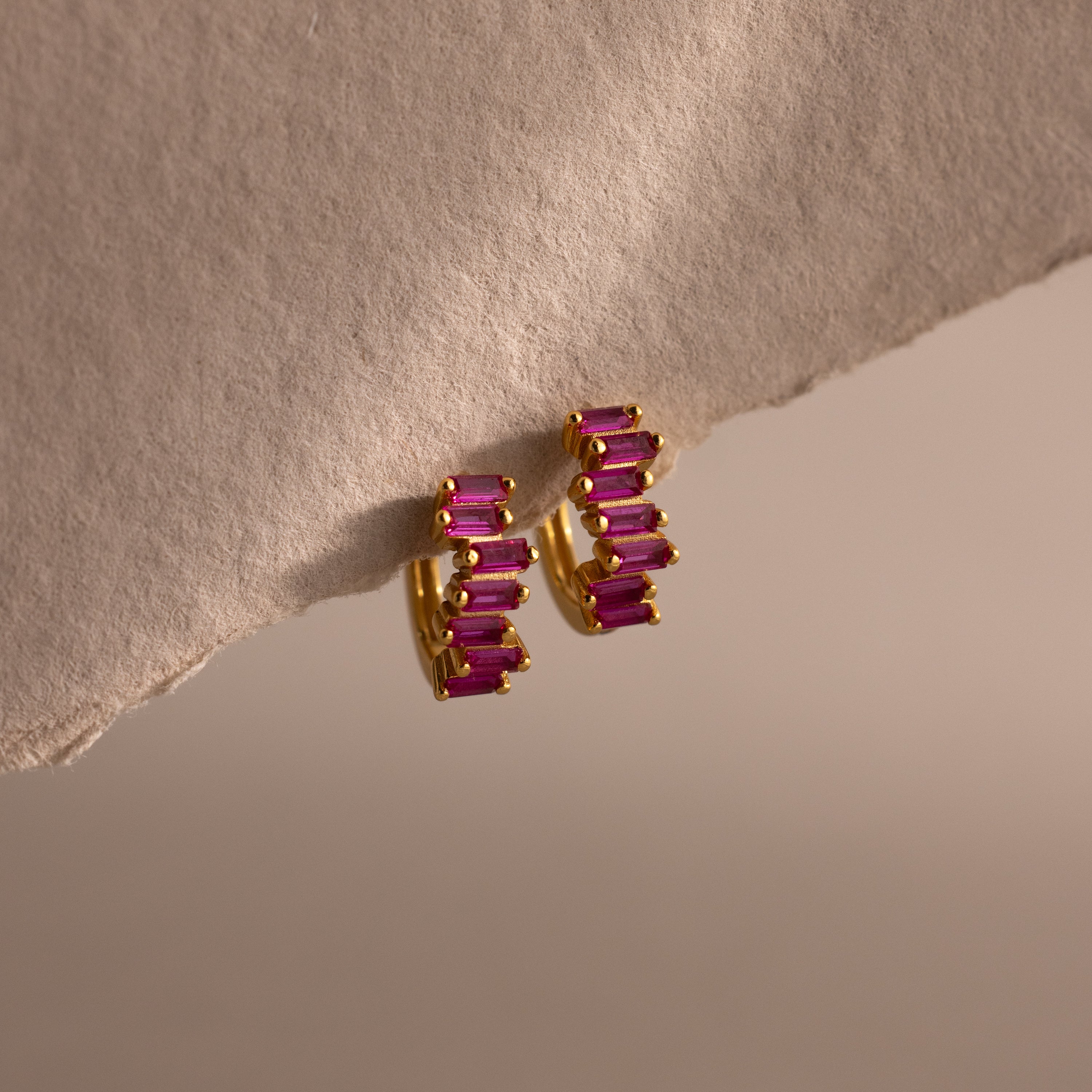 Staggered Baguette Ruby Huggies, featuring gold hoops with rectangular pink baguette-cut stones, are displayed on a textured neutral surface under soft lighting for a modern, elegant touch.