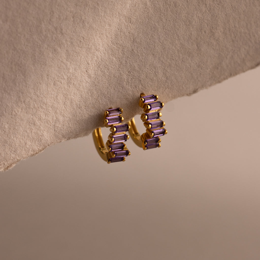 The Staggered Baguette Amethyst Huggies are modern gold hoops adorned with three rectangular amethyst stones on each, elegantly displayed on a textured beige surface.