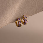 The Staggered Baguette Amethyst Huggies are modern gold hoops adorned with three rectangular amethyst stones on each, elegantly displayed on a textured beige surface.