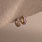 Staggered Baguette Alexandrite Huggies—gold modern hoop earrings with three baguette-cut purple stones—displayed on a textured beige surface.