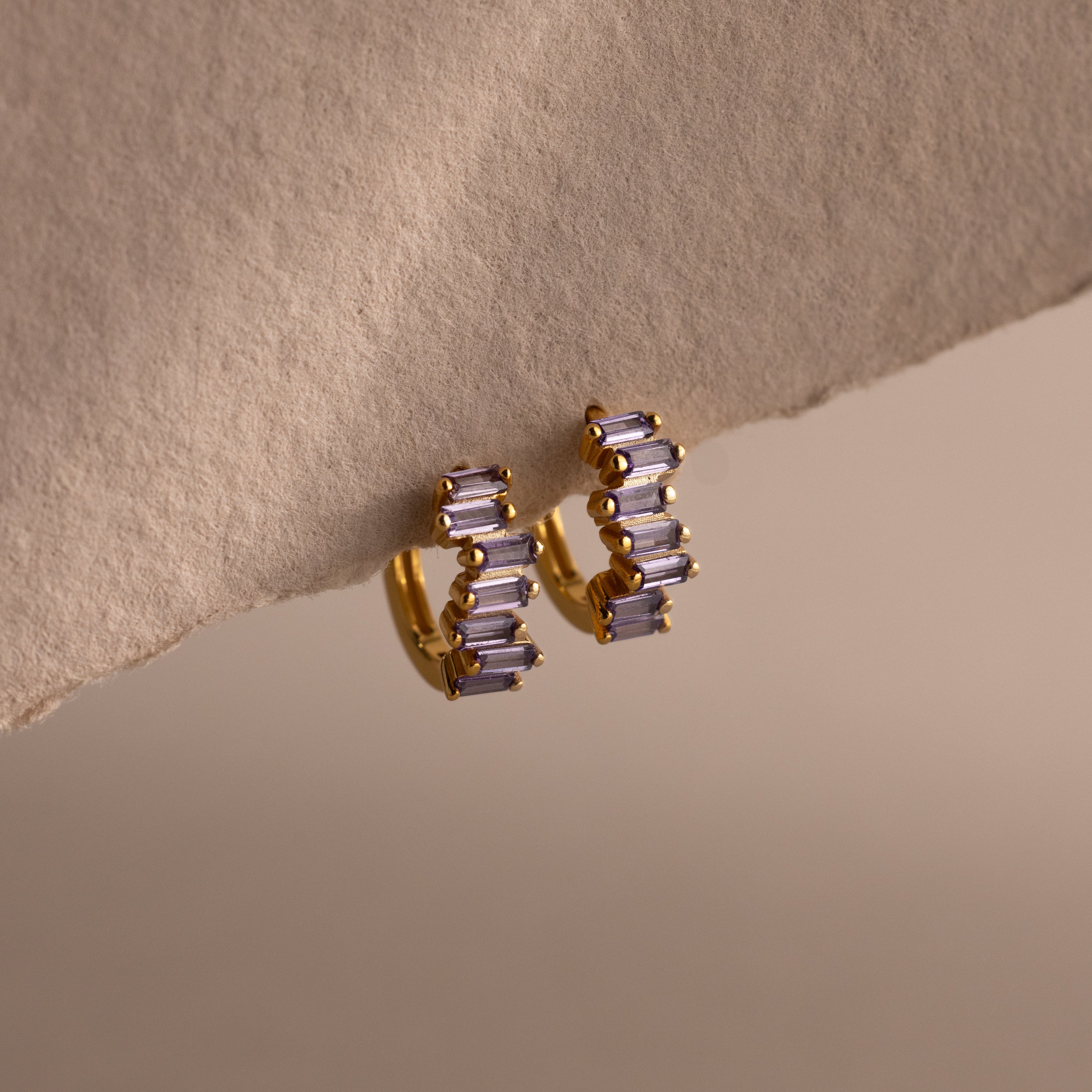 Staggered Baguette Alexandrite Huggies—gold modern hoop earrings with three baguette-cut purple stones—displayed on a textured beige surface.