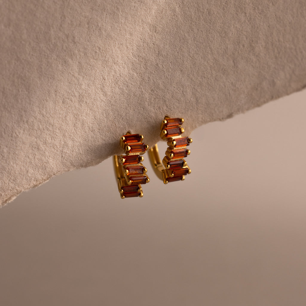 Staggered Baguette Garnet Huggies, featuring rectangular red stones, display modern elegance against a textured beige background.