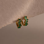 Staggered Baguette Emerald Huggies showcase modern elegance with vertically stacked rectangular green stones, their baguette-cut emeralds adding striking detail to any look on a textured beige surface.