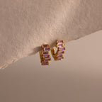 Staggered Baguette Pink Tourmaline Huggies—gold earrings each with three baguette-cut pink tourmaline stones—displayed on a textured beige surface, capturing modern elegance in every detail.