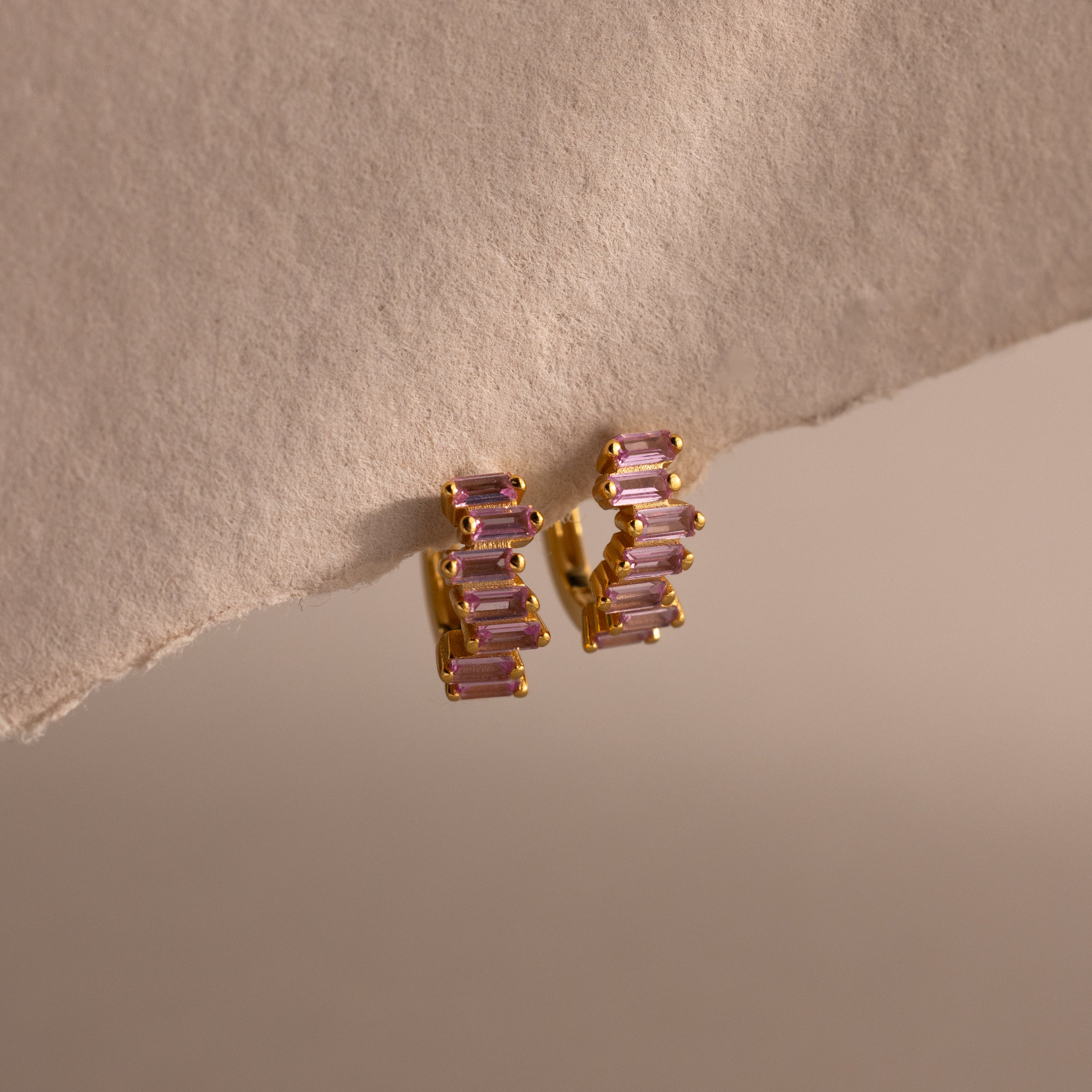 Staggered Baguette Pink Tourmaline Huggies—gold earrings each with three baguette-cut pink tourmaline stones—displayed on a textured beige surface, capturing modern elegance in every detail.
