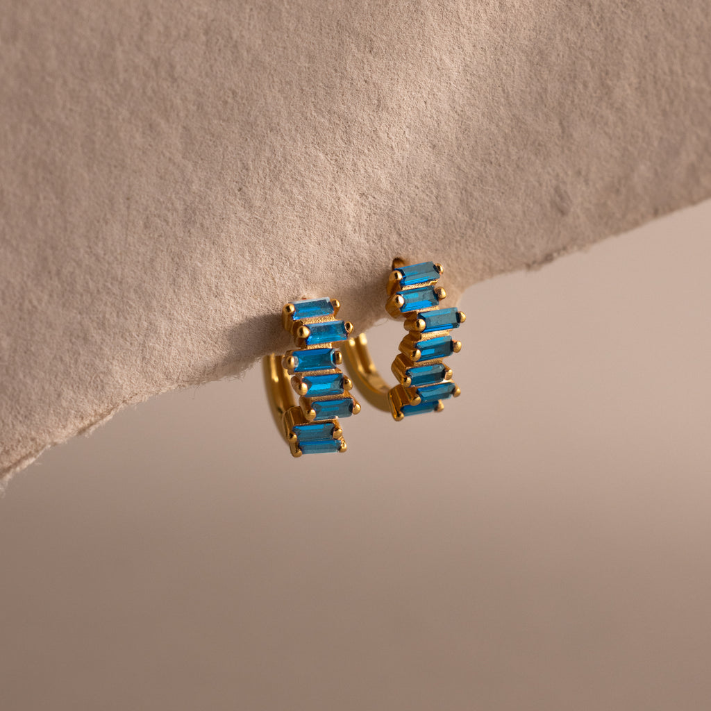 Staggered Baguette Blue Zircon Huggies displayed on a textured beige surface, highlighting modern elegance with their gold hoops and baguette-cut blue zircon stones.