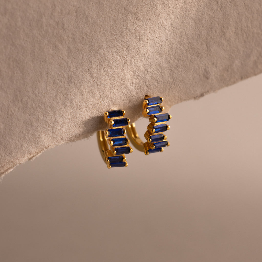 Staggered Baguette Sapphire Huggies with rectangular blue baguette-cut stones, shown hanging from a textured, beige surface.