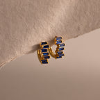 Staggered Baguette Sapphire Huggies with rectangular blue baguette-cut stones, shown hanging from a textured, beige surface.