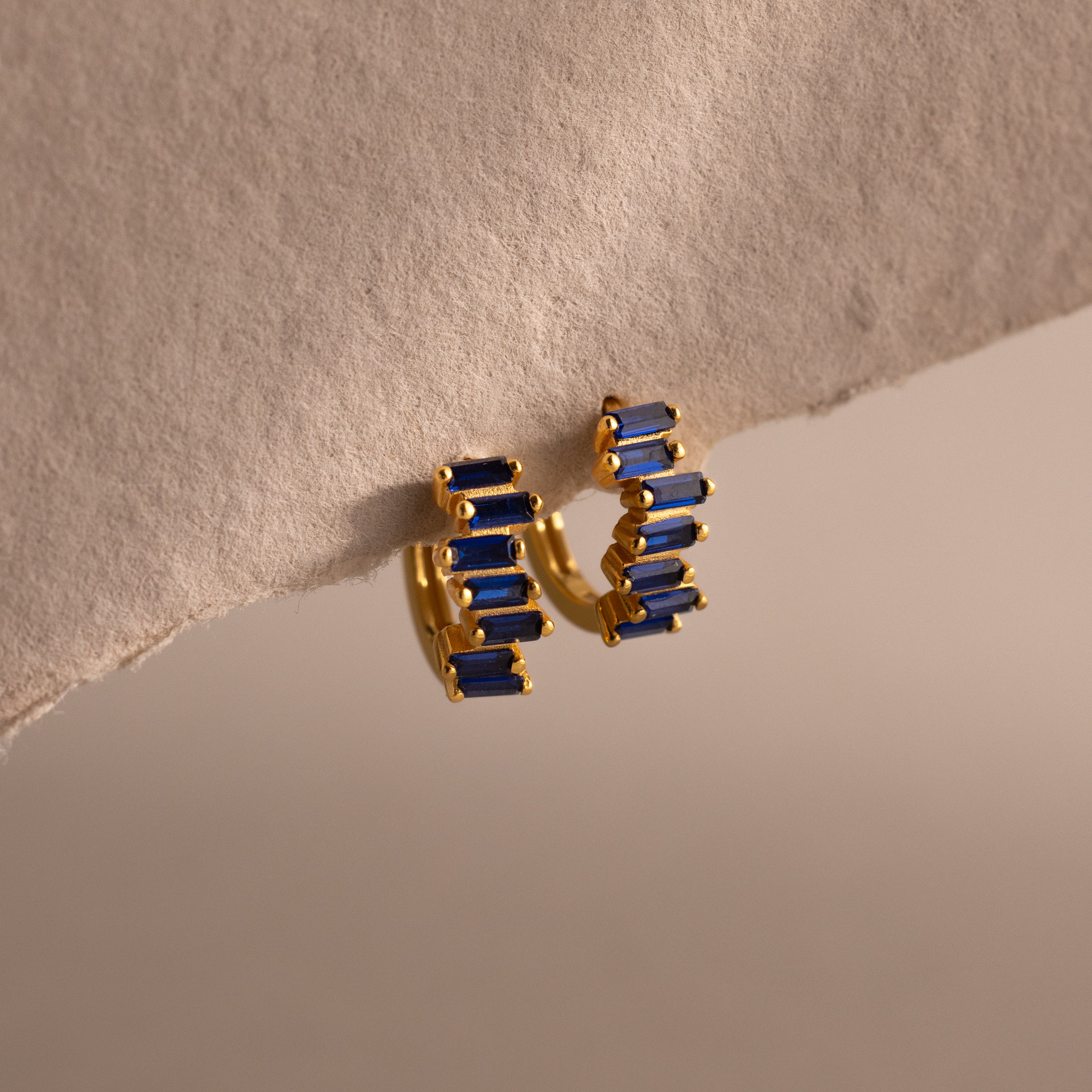Staggered Baguette Sapphire Huggies with rectangular blue baguette-cut stones, shown hanging from a textured, beige surface.