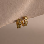 The Staggered Baguette Peridot Huggies exude modern elegance, featuring baguette-cut peridots in a gold setting and displayed on the edge of a textured beige surface.