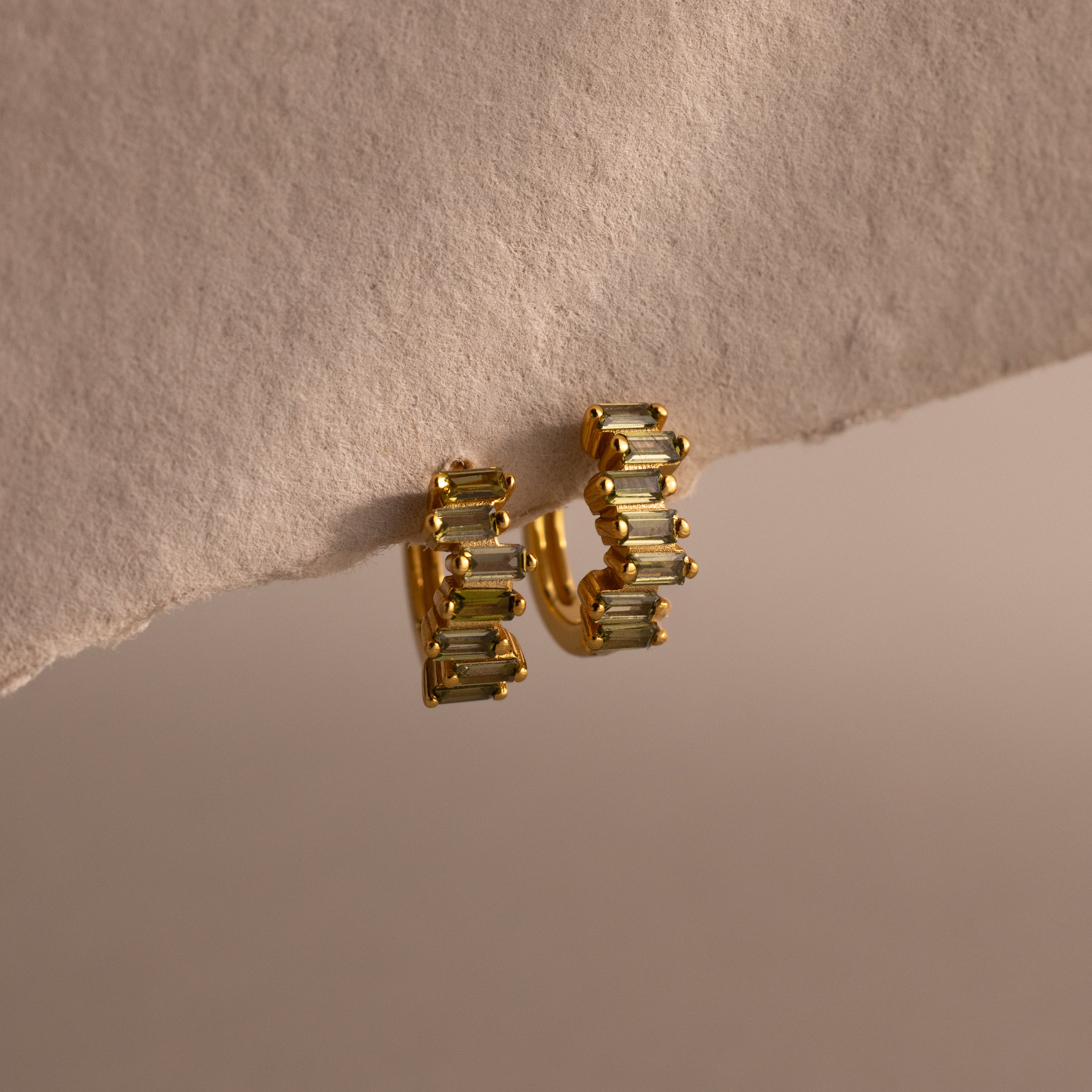 The Staggered Baguette Peridot Huggies exude modern elegance, featuring baguette-cut peridots in a gold setting and displayed on the edge of a textured beige surface.