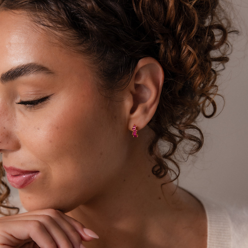 A woman with curly hair wears the Staggered Baguette Birthstone Huggies, embodying modern elegance as she rests her chin on her hand and looks down.