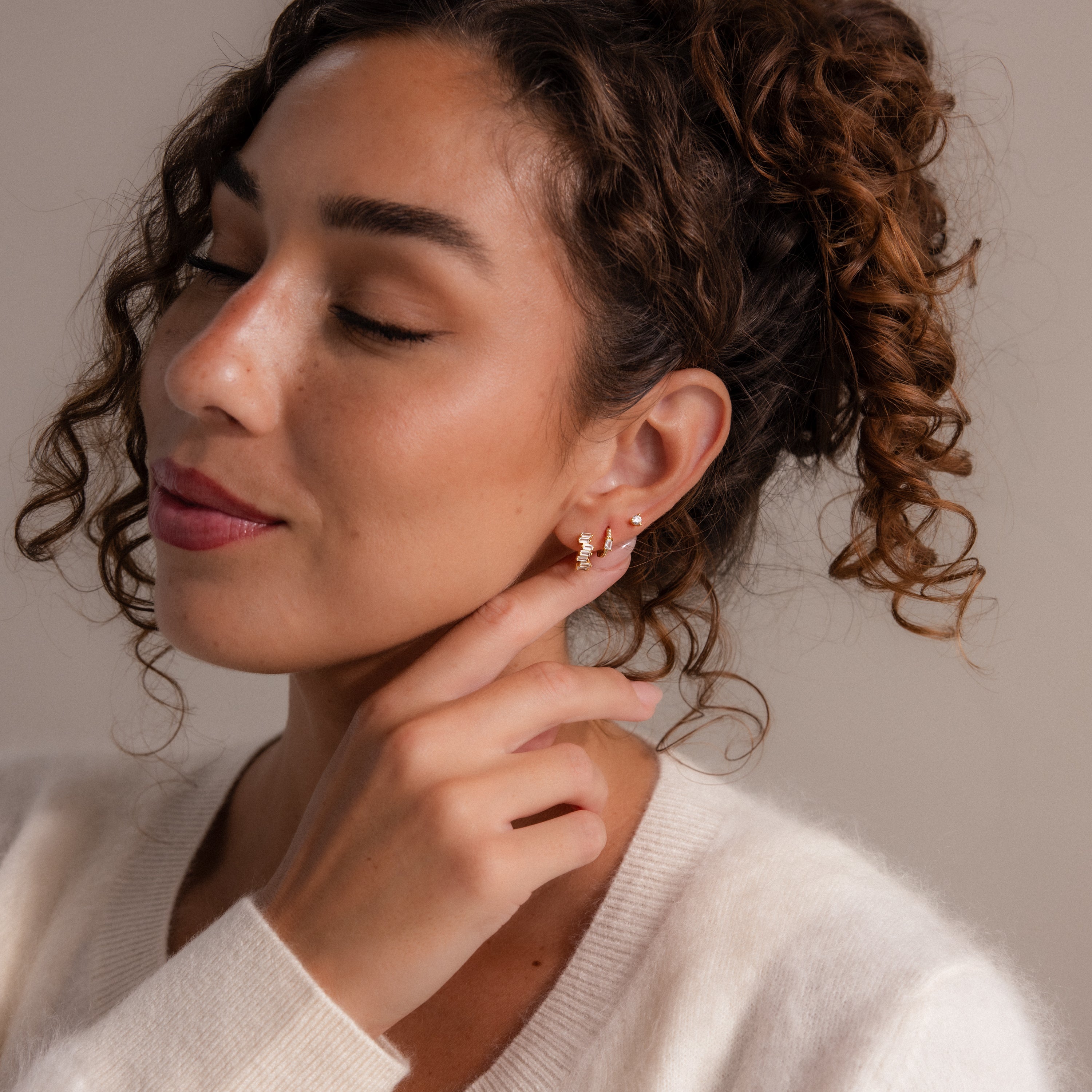 A woman with curly hair smiles gently, touching her Staggered Baguette Birthstone Huggies and radiates modern elegance in a cozy white sweater.