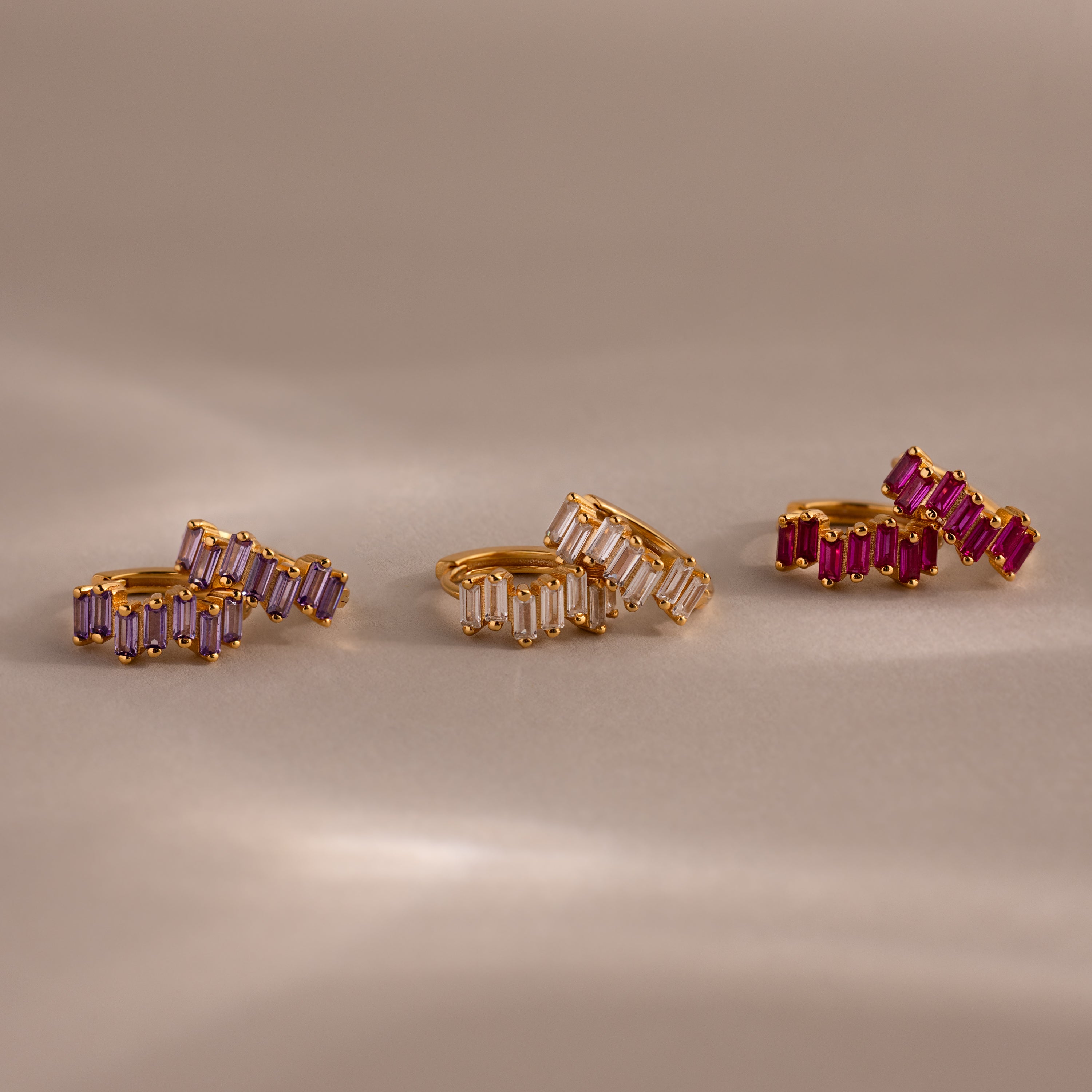 Three pairs of Staggered Baguette Garnet Huggies with rectangular gemstones in purple, clear, and pink shades showcase modern elegance on a beige surface.