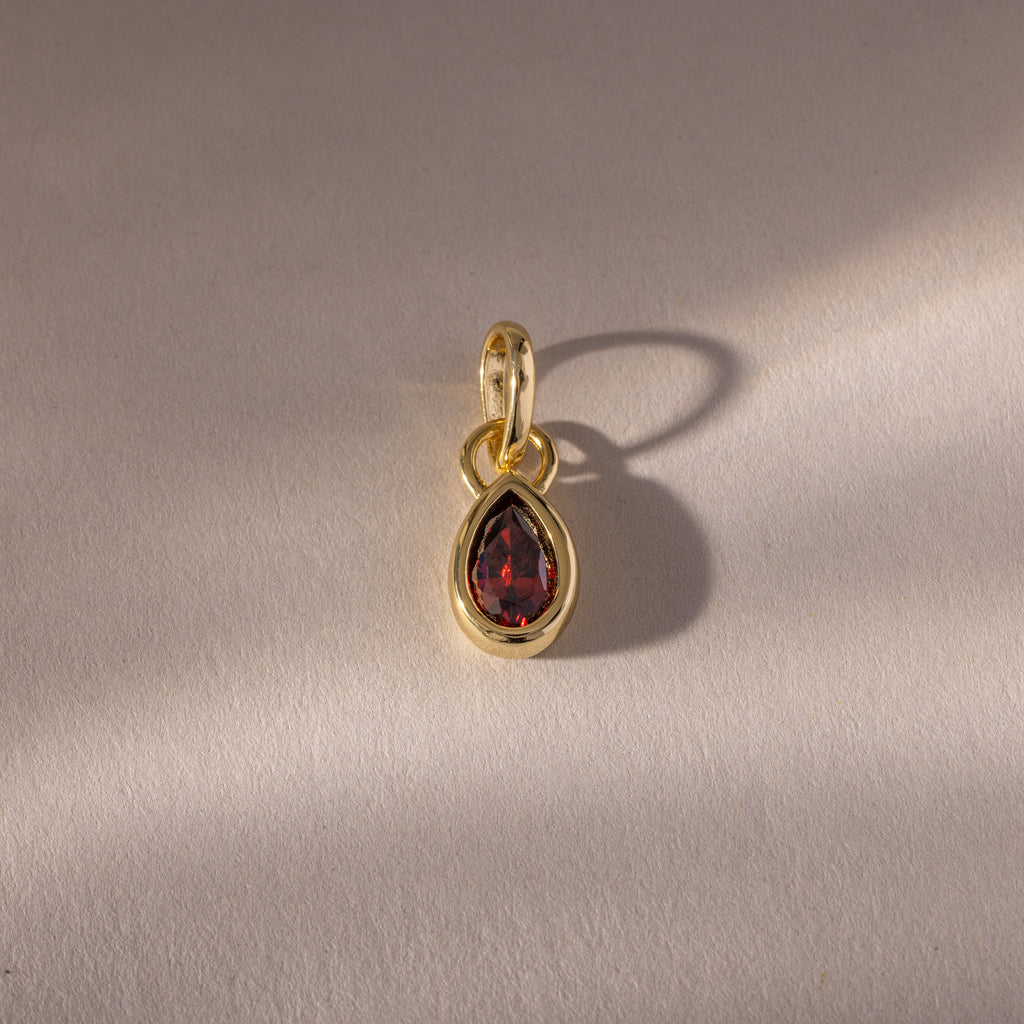 The Teardrop Garnet Charm features a teardrop-shaped red gemstone in a gold bezel setting, casting a soft shadow on a textured beige surface.