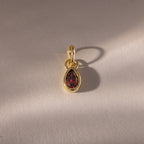 The Teardrop Garnet Charm features a teardrop-shaped red gemstone in a gold bezel setting, casting a soft shadow on a textured beige surface.