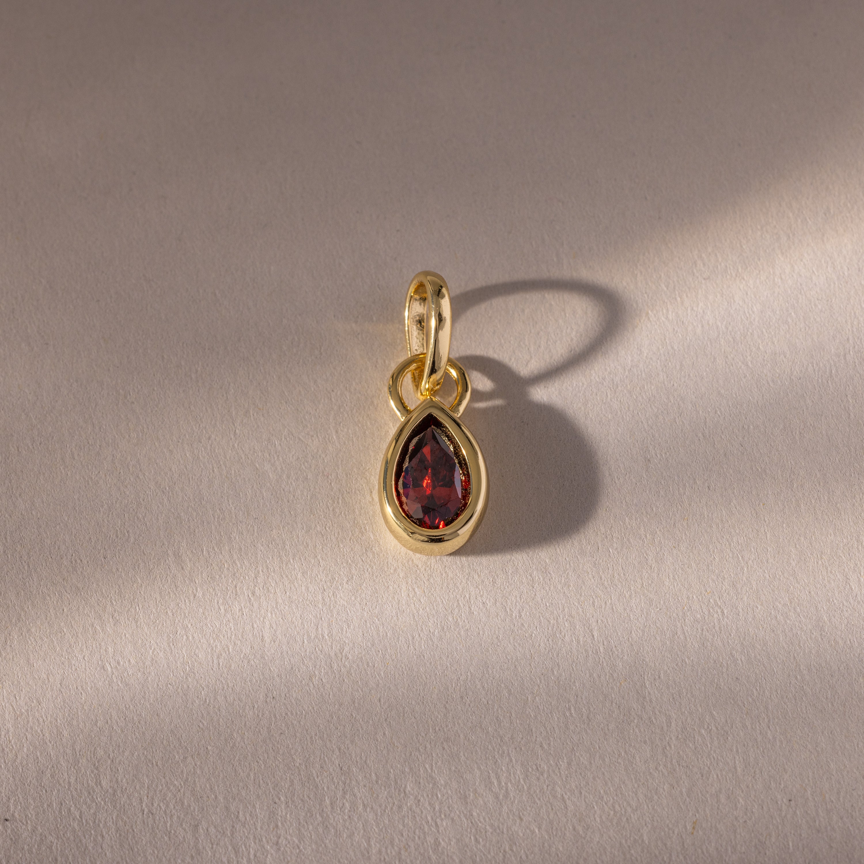 The Teardrop Garnet Charm features a teardrop-shaped red gemstone in a gold bezel setting, casting a soft shadow on a textured beige surface.