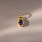 The Teardrop Amethyst Charm features a purple teardrop-shaped gemstone in a gold bezel setting, displayed on a light textured surface and casting a soft shadow.