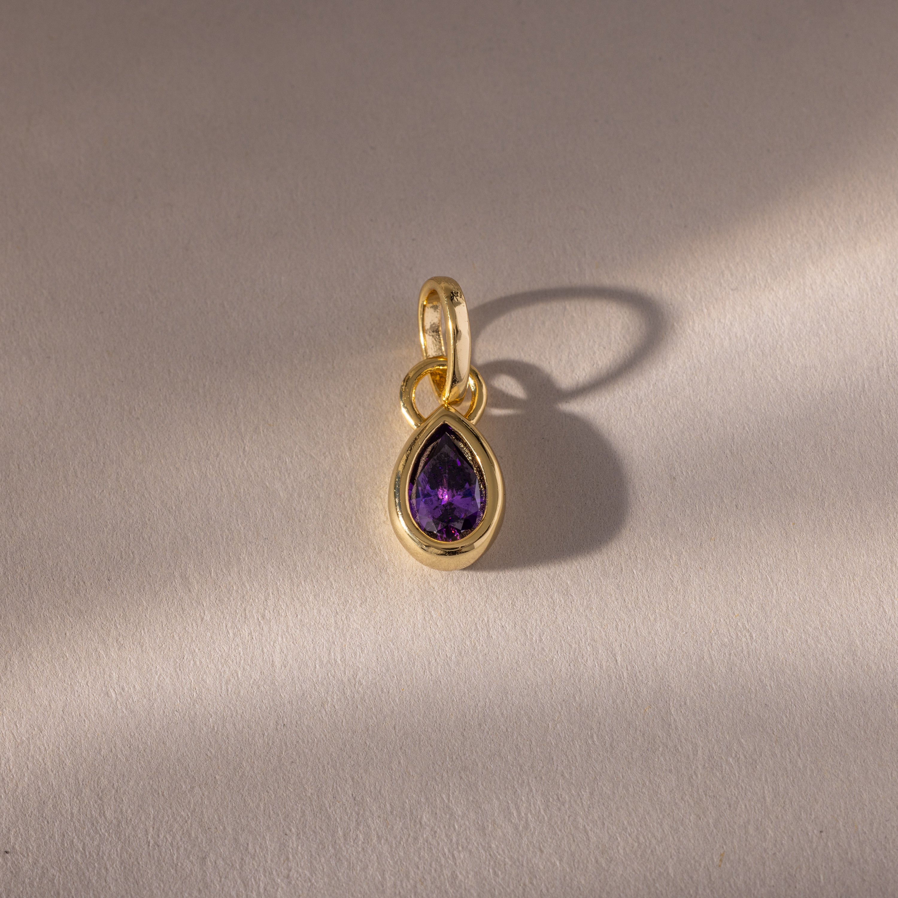 The Teardrop Amethyst Charm features a purple teardrop-shaped gemstone in a gold bezel setting, displayed on a light textured surface and casting a soft shadow.