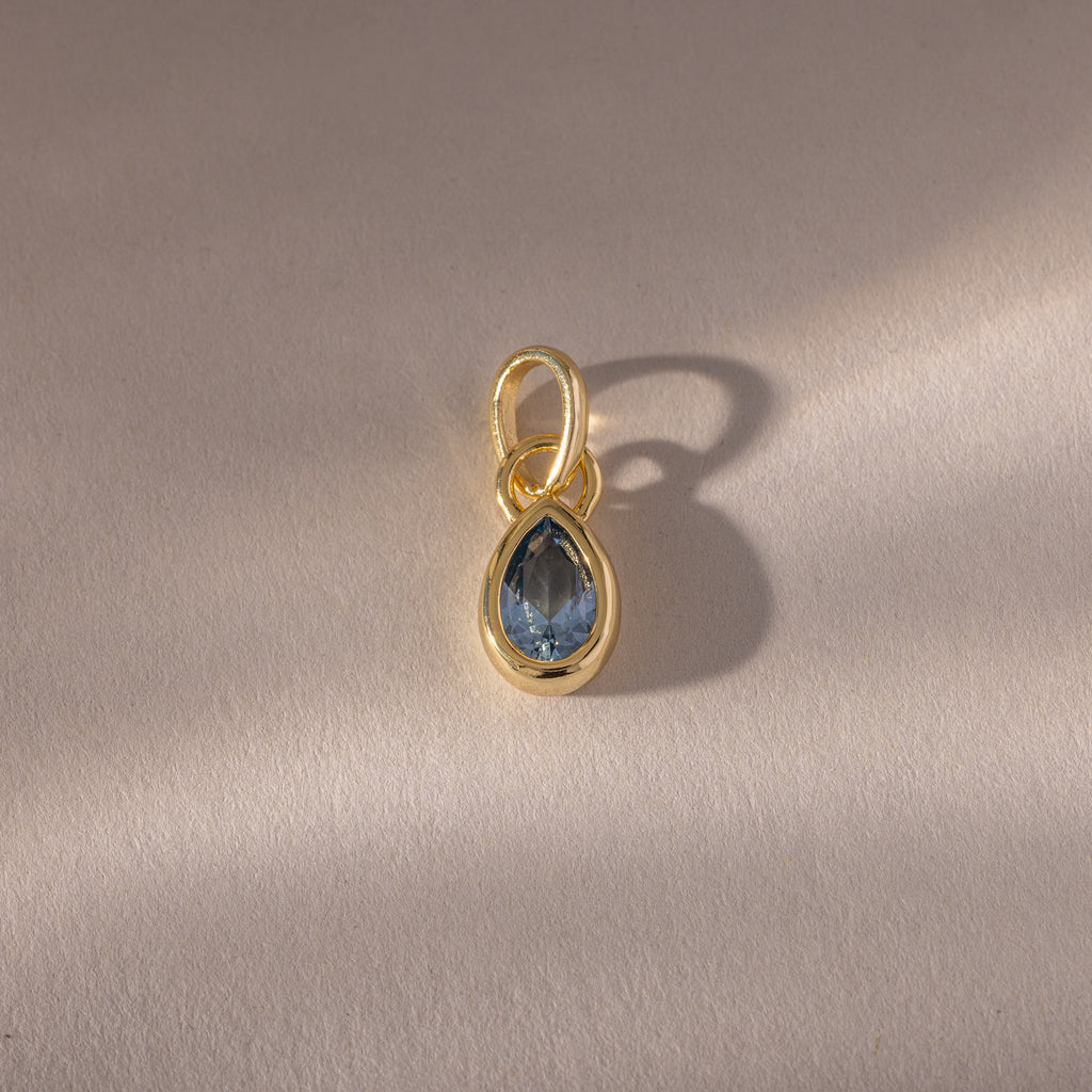 The Teardrop Aquamarine Charm features a blue teardrop birthstone in a gold bezel setting, showcased on a light beige background.