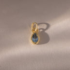 The Teardrop Aquamarine Charm features a blue teardrop birthstone in a gold bezel setting, showcased on a light beige background.
