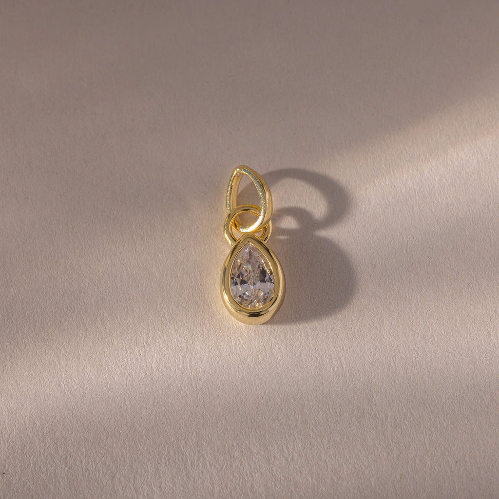The Teardrop Diamond Charm features a gold teardrop design with a clear gemstone in a gold bezel setting, displayed on a light textured background with a soft shadow.
