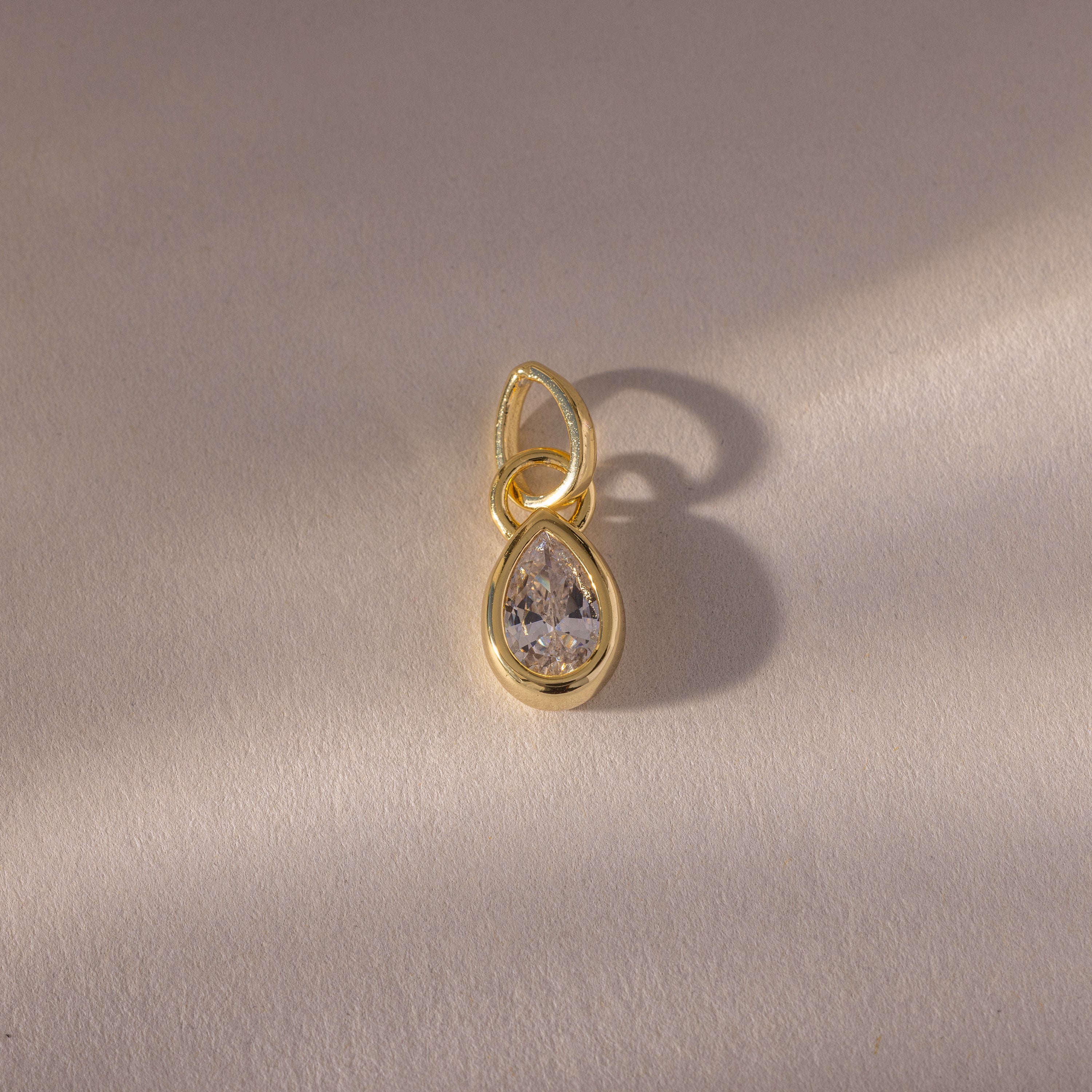 The Teardrop Diamond Charm features a gold teardrop design with a clear gemstone in a gold bezel setting, displayed on a light textured background with a soft shadow.
