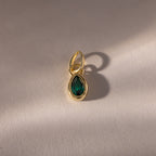 The Teardrop Emerald Charm features a teardrop-shaped green gemstone in a gold bezel setting, casting a soft shadow on a light surface—a timeless piece of jewelry.
