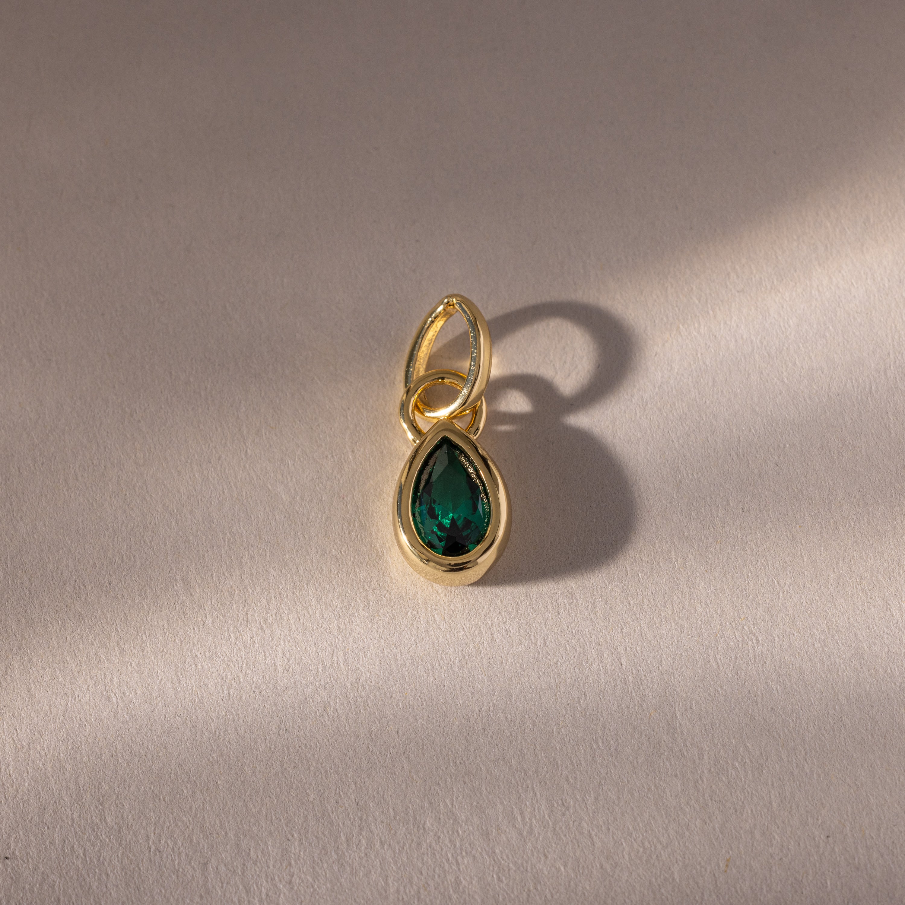 The Teardrop Emerald Charm features a teardrop-shaped green gemstone in a gold bezel setting, casting a soft shadow on a light surface—a timeless piece of jewelry.