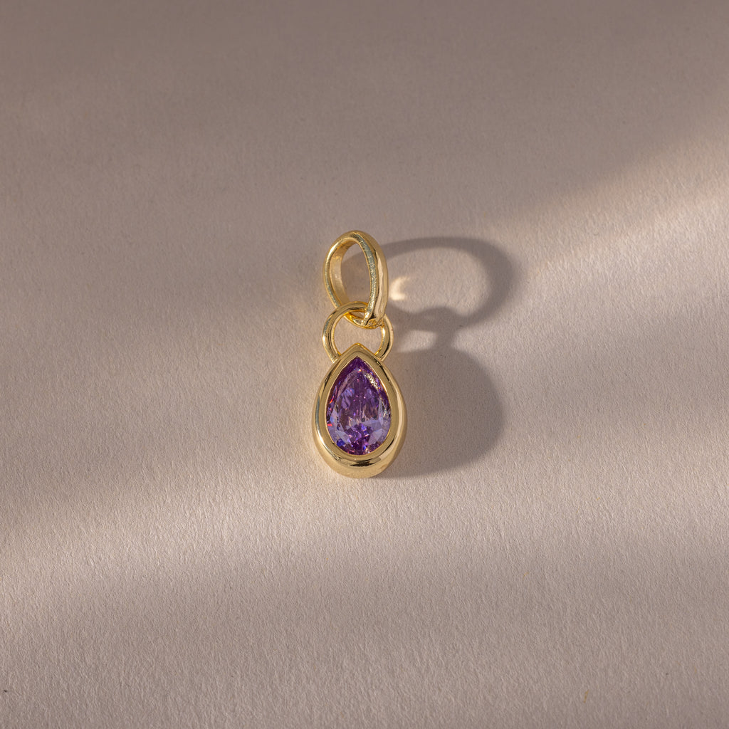 A close-up of the Teardrop Alexandrite Charm reveals its intricate gemstone set in a gold bezel setting.