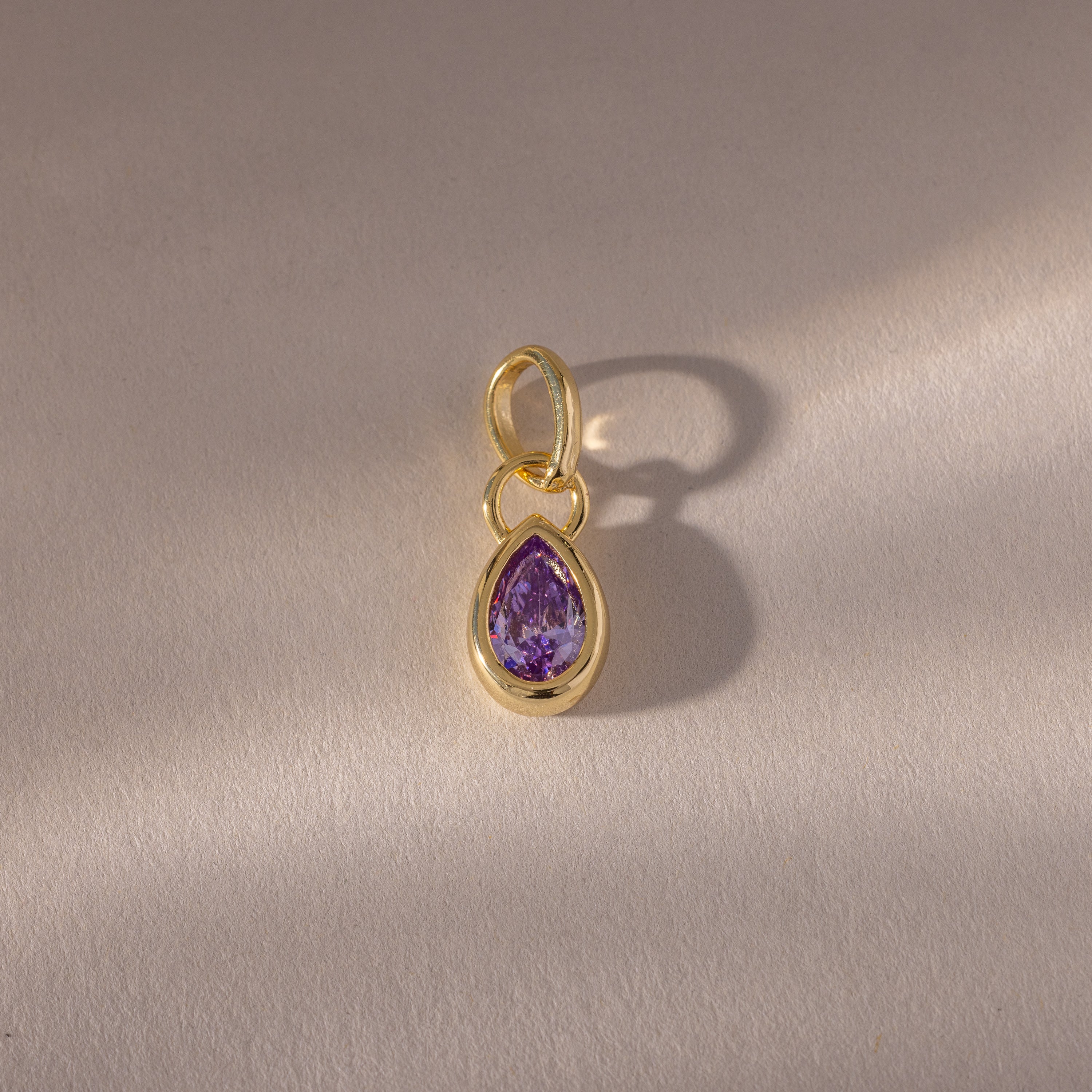A close-up of the Teardrop Alexandrite Charm reveals its intricate gemstone set in a gold bezel setting.
