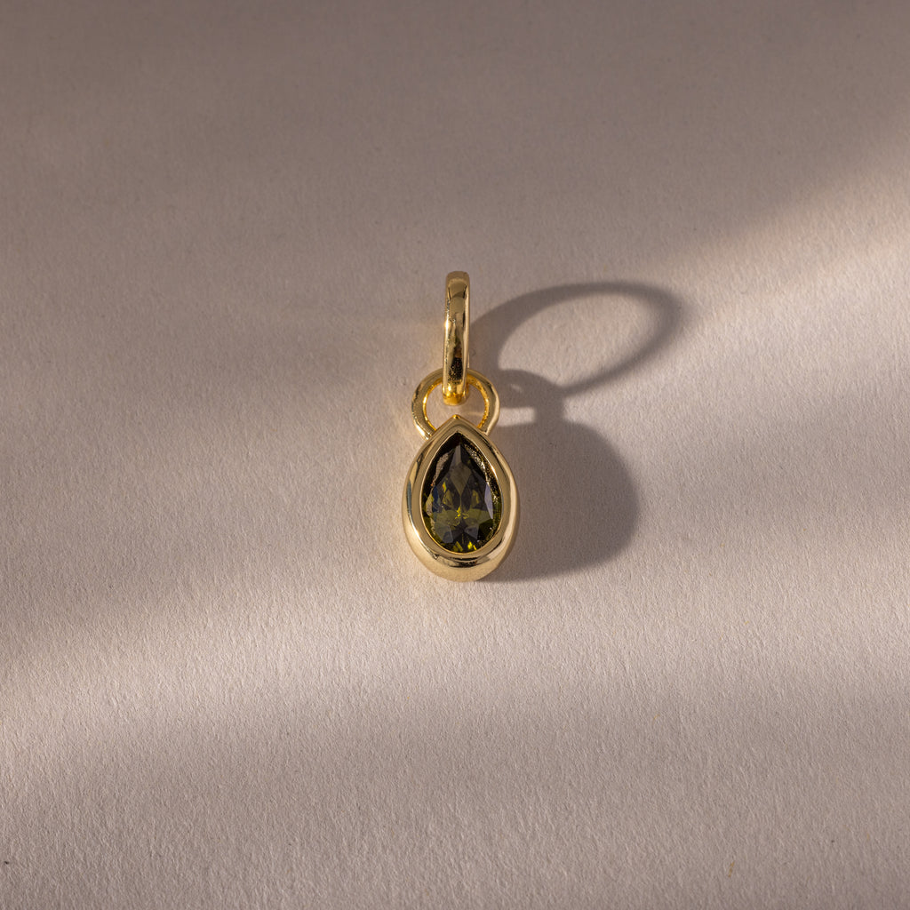The Teardrop Peridot Charm features a vibrant green peridot in a gold bezel setting, its gemstone casting a subtle shadow—an elegant addition to any jewelry collection.