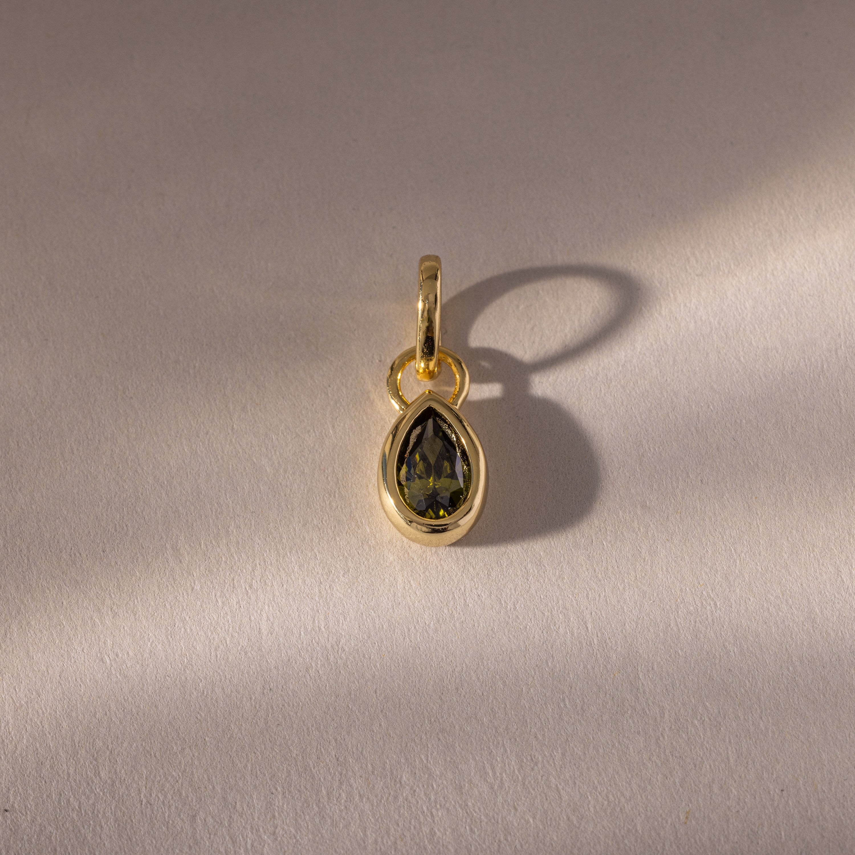 The Teardrop Peridot Charm features a vibrant green peridot in a gold bezel setting, its gemstone casting a subtle shadow—an elegant addition to any jewelry collection.