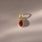 The Teardrop Ruby Charm features a teardrop-shaped red gemstone set in a gold bezel, casting a delicate shadow on a textured surface—a refined piece of jewelry.