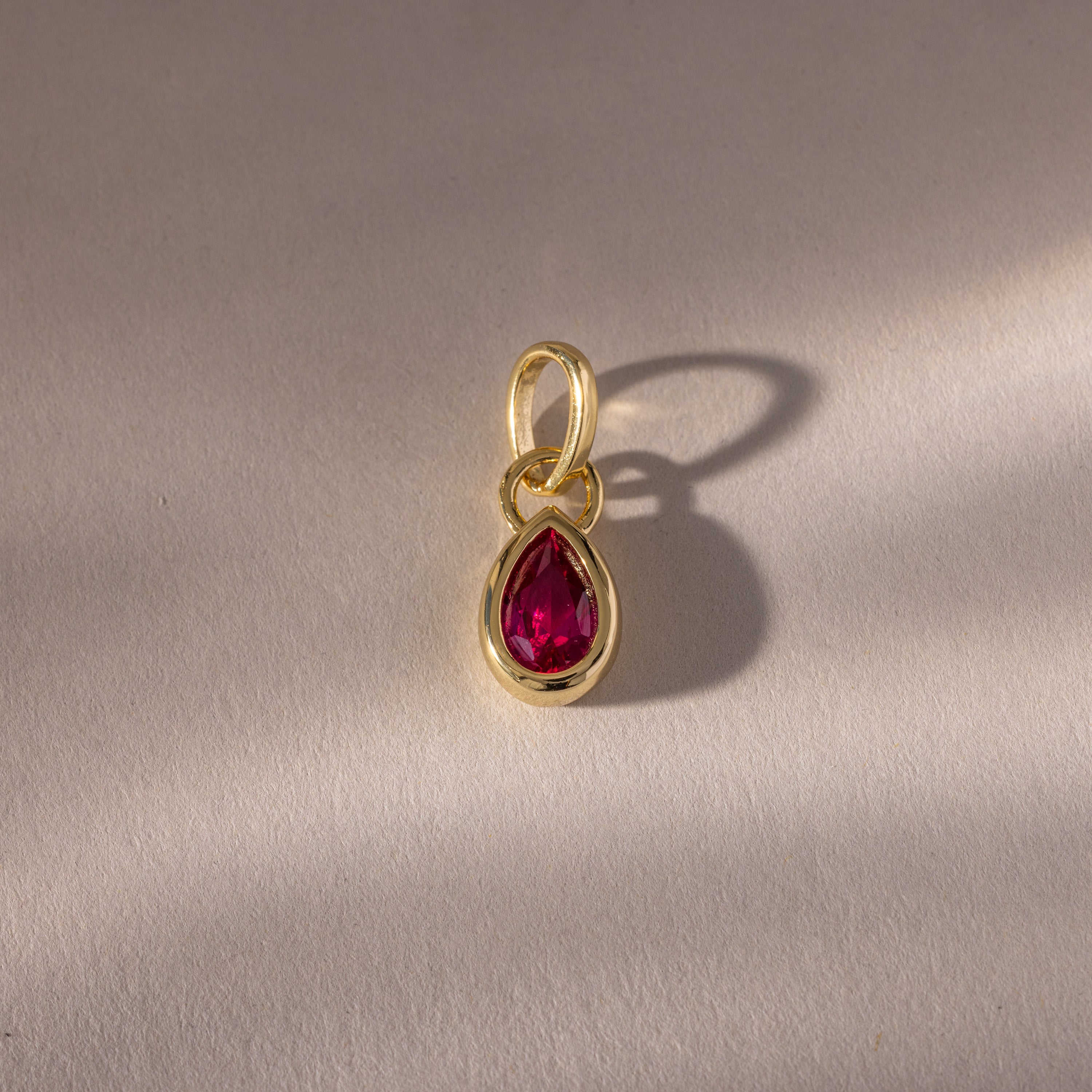 The Teardrop Ruby Charm features a teardrop-shaped red gemstone set in a gold bezel, casting a delicate shadow on a textured surface—a refined piece of jewelry.