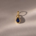 The Teardrop Sapphire Charm is a gold pendant with a teardrop-shaped sapphire in a bezel setting, featuring a blue gemstone that casts a shadow on a light textured surface.